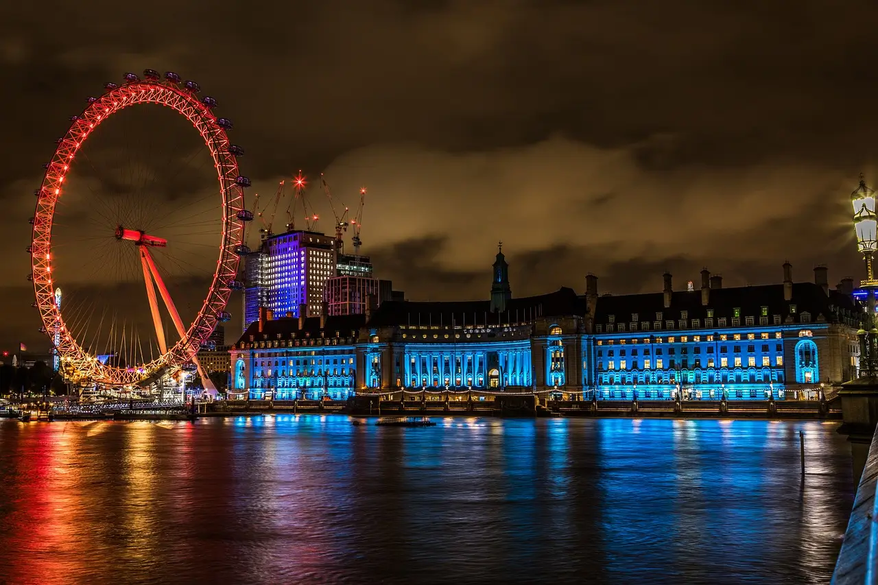 The London Eye illuminated in purple and blue, reflecting in the River Thames at night during the London By Night Luxury Chauffeured Tour.